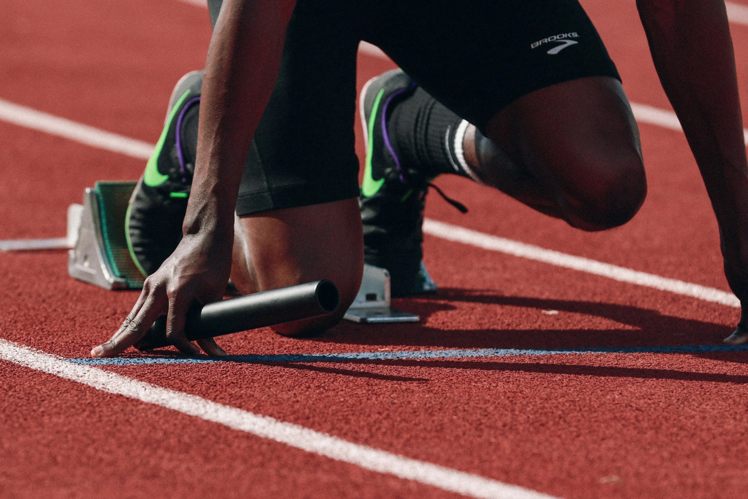 Close-up of a male athlete in a starting position on a red running track, gripping the starting block handle with his left hand, dressed in black sportswear and running shoes.